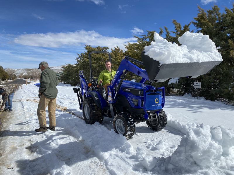 A man operating a blue tractor with the front end loader full of snow along a snowy street.