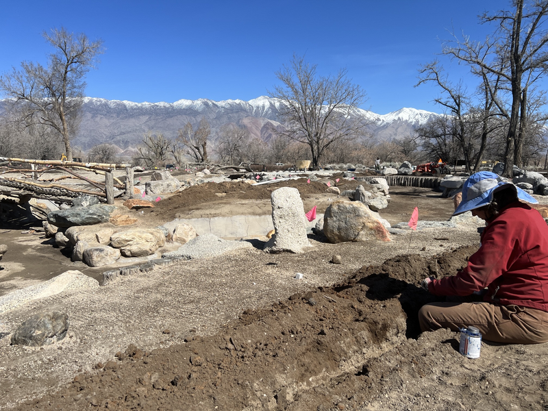 A woman installing water pipe and sprinklers in a dry rock garden with a bridge and snowy mountains in the background.