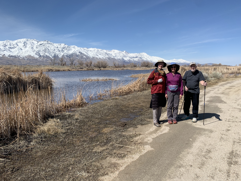 3 people standing next to a pond with snowy mountains behind