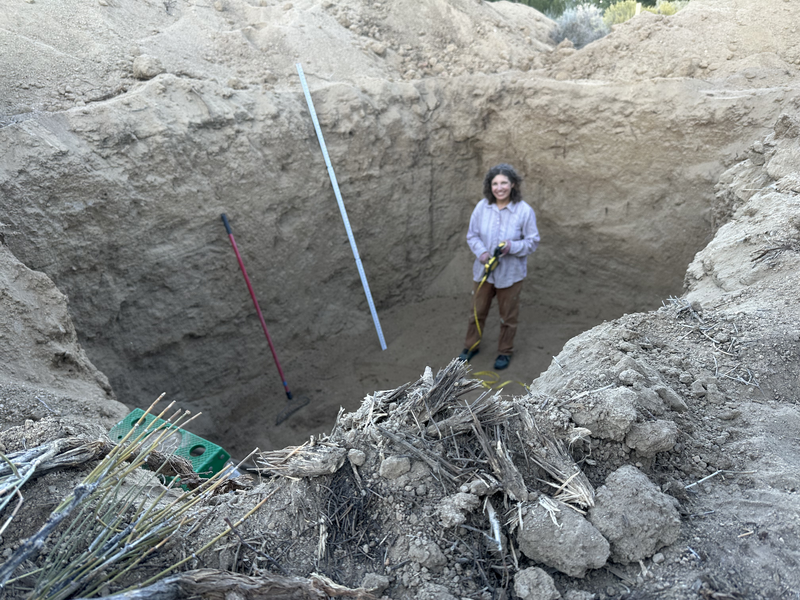 A smiling woman in a large hole with a zip level and a rake