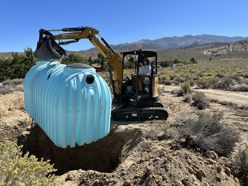 A woman lowers a large blue septic tank into a hole using a mini excavator