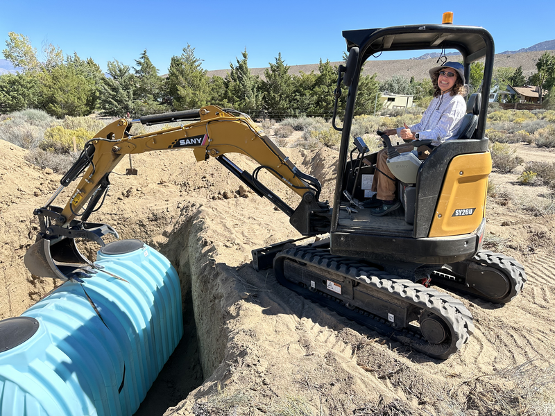 A woman in a mini-excavator with a large blue septic tank in a large hole