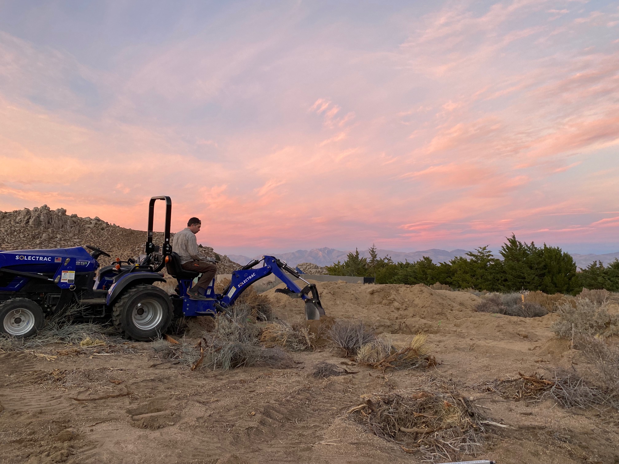 A man using a backhoe attachment at pink glowing sunset