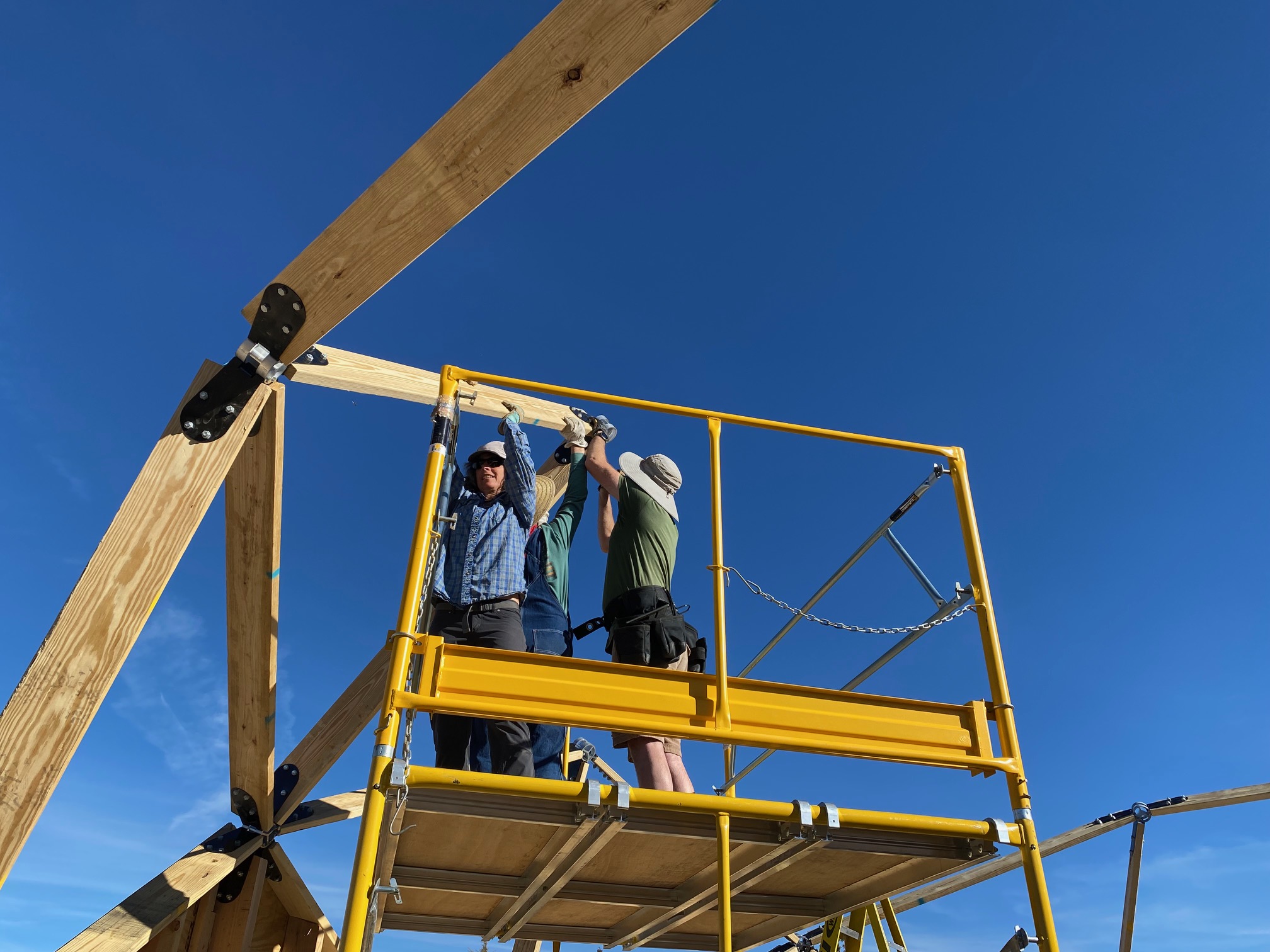 Two people hold struts up, while a third nails a pin in the hub connecting them, as part of geodesic dome construction.