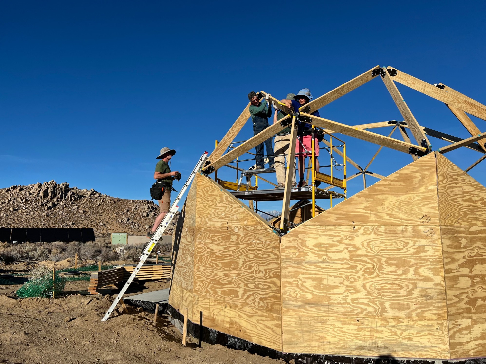 3 people crowd the scaffolding and 1 is on the extension ladder to fit and connect 2 stubborn struts on a geodesic dome