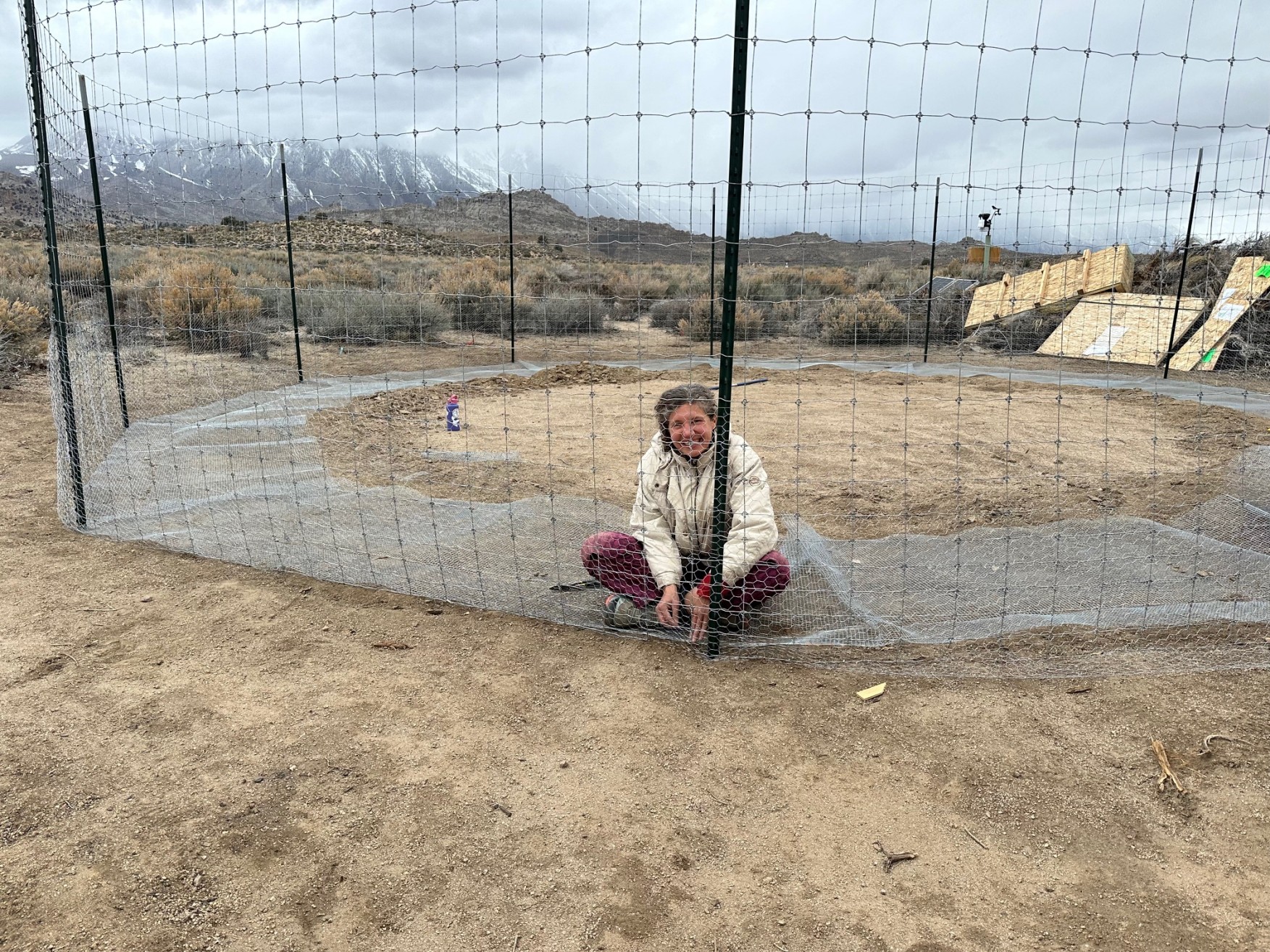 A woman inside a fenced garden, sitting on some hardware cloth.