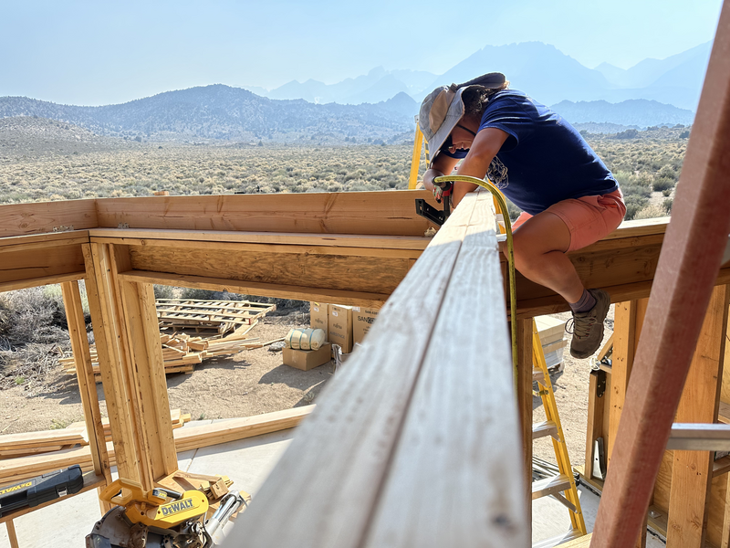 A woman sitting on a wall, using an air nailer on a ledger board