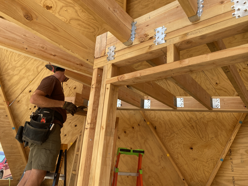 A man measures in a corner, but in the foreground are joists held in place with shiny metal hangers 